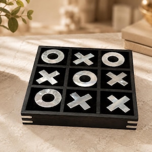 May include: A square black wooden tic-tac-toe game board featuring a grid of nine compartments. The game includes iridescent white shell-like X and O playing pieces arranged within the slots. The board has small silver-toned metal accents on corners.