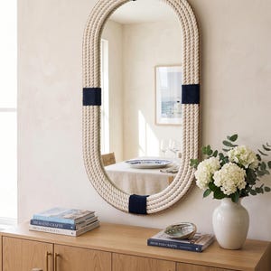 May include: Oval mirror with a thick, off-white rope frame, accented with navy blue bands. The mirror hangs on a light beige wall above a wooden cabinet. A vase of white flowers and books are on the cabinet.