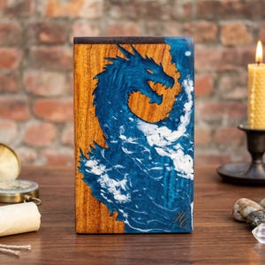 May include: Wooden box with a blue dragon design. The dragon is set against a wood grain background with white accents. The box is rectangular and has a dark brown border. A lit candle and other decorative items are in the background.