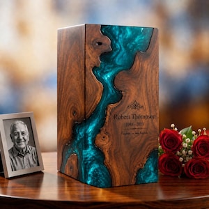 May include: A rectangular wooden cremation urn featuring a central river-like inlay of translucent teal resin. The wood has a dark brown grain. Engraved text reads: Robert Thompson, 1948-2023, Forever in our hearts, with a decorative flourish.