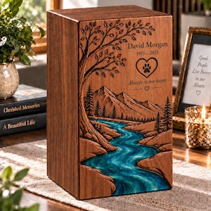 May include: A rectangular wooden cremation urn featuring a laser-engraved landscape of a tree, mountains, and a forest. The river is filled with blue resin. The text reads David Morgan 1955-2023, Always in our hearts, and a heart with a paw print.
