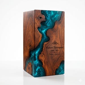 May include: A rectangular wooden cremation urn featuring a natural dark brown wood grain and a central flowing inlay of translucent teal blue resin. The front surface is engraved with the text Robert Thompson, 1948-2023, and Forever in our hearts.