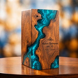 May include: A rectangular wooden cremation urn featuring a vibrant teal resin inlay that mimics a flowing river. The wood has a natural grain pattern, and the front is engraved with the text Robert Thompson, 1948-2023, and Forever in our hearts.