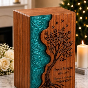 May include: A rectangular wooden cremation urn featuring a vertical, wavy inlay of translucent teal resin. The wood surface is engraved with a detailed tree, birds, and the text: David Morgan, 1955-2023, Forever in our hearts.