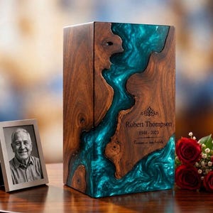 May include: A rectangular wooden cremation urn featuring a central flowing inlay of swirling teal resin. The wood has a natural grain pattern. Engraved text reads: Robert Thompson, 1948-2023, and Forever in our hearts, with a decorative flourish.