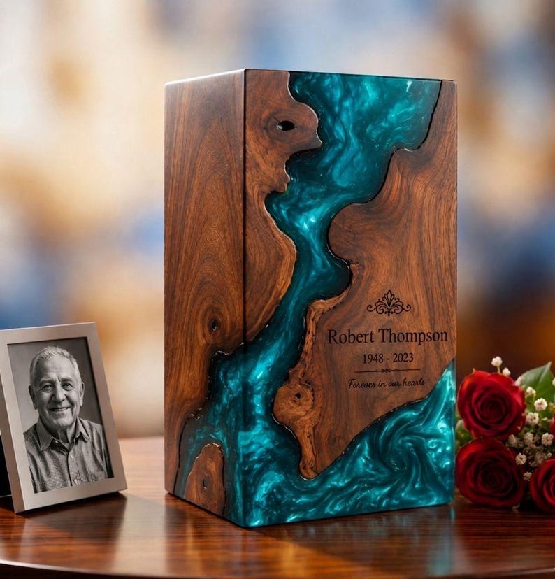 May include: A rectangular wooden cremation urn featuring a central flowing inlay of swirling teal resin. The wood has a natural grain pattern. Engraved text reads: Robert Thompson, 1948-2023, and Forever in our hearts, with a decorative flourish.