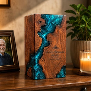 May include: A rectangular wooden cremation urn featuring a central river-like inlay of translucent teal epoxy resin. The front surface is engraved with the name Robert Thompson, the years 1948-2023, and the phrase Forever in our hearts.