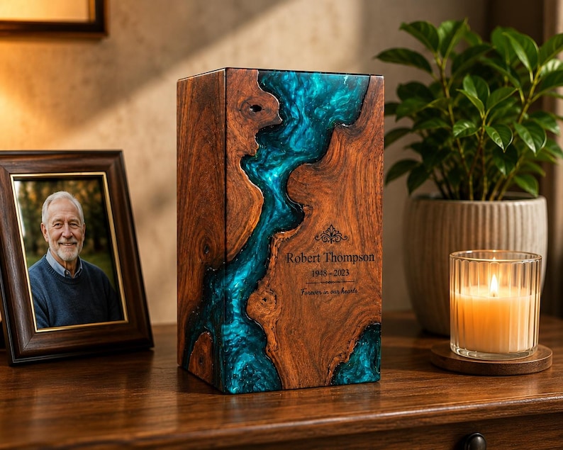 May include: A rectangular wooden cremation urn featuring a central river-like inlay of translucent teal epoxy resin. The front surface is engraved with the name Robert Thompson, the years 1948-2023, and the phrase Forever in our hearts.