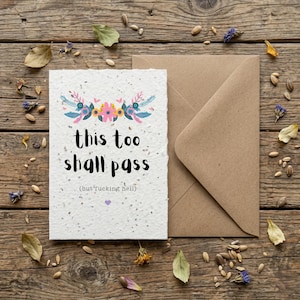 May include: A cream-coloured greeting card with the words "this too shall pass" and the phrase "(but f*cking hell)". The card features a floral design and a brown envelope, on a wooden surface with seeds and dried flowers scattered around.