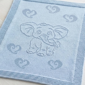 May include: A light blue knitted blanket featuring a central elephant motif surrounded by five stylized heart shapes. The rectangular blanket has a textured garter stitch border framing the smooth stockinette stitch center panel with the design.