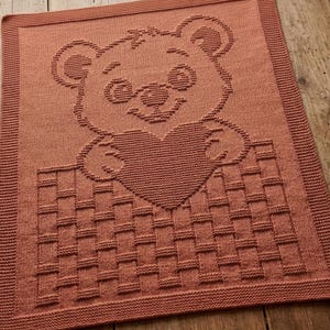 May include: A rectangular terracotta-coloured knitted blanket featuring a textured design of a teddy bear holding a heart. The bear is positioned above a woven basket pattern, and the entire piece is framed by a simple garter stitch border.