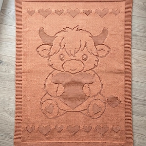May include: A rectangular, peach-coloured knitted blanket featuring a central textured design of a cartoon highland cow holding a large heart. The border is framed by a garter stitch pattern, with rows of small knitted hearts above and below the cow.