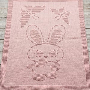 May include: A rectangular light pink knitted blanket featuring a textured pattern of a central rabbit character with large ears and two butterflies positioned above the rabbit. The blanket has a wide, uniform garter stitch border around the edges.