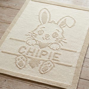 May include: A cream-coloured knitted blanket featuring a textured design of a rabbit peeking over a horizontal line. Below the line, the name CHIPIE is knitted in capital letters, flanked by two paw prints and a heart shape, with a ribbed border.