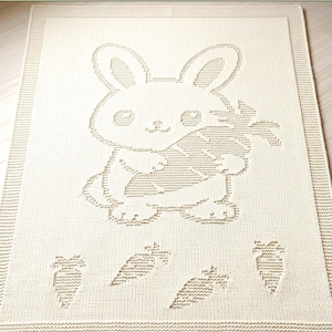 K&ouml;nnte beinhalten: Eine rechteckige, cremefarbene Strickdecke mit einem strukturierten Design eines Hasen, der eine gro&szlig;e Karotte h&auml;lt. Der Hase ist mittig platziert, darunter befinden sich vier kleinere Karottenmotive in einer horizontalen Reihe. Breiter Rand.