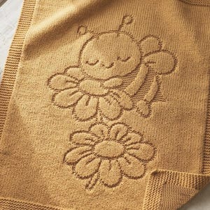 May include: A rectangular mustard yellow knitted blanket featuring a textured relief design of a sleeping bee resting on a flower above a second standalone flower. The blanket has a wide garter stitch border framing the central knit pattern.