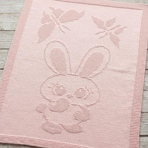 May include: A rectangular light pink knitted blanket featuring a textured pattern of a central rabbit character and two butterflies above it. The blanket has a wide, uniform knitted border framing the central design on all four sides.