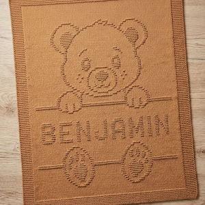 May include: A rectangular, tan-colored knitted blanket featuring a textured, raised pattern of a teddy bear peeking over a horizontal line. Below the bear, the name BENJAMIN is knitted in capital letters, followed by two bear paw prints.