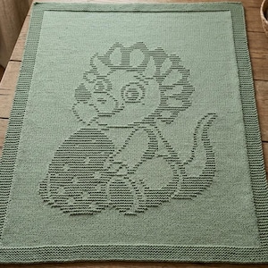 Puede incluir: Una manta de punto rectangular de color verde salvia con un diseño de intarsia monocromático y texturizado de un dinosaurio con una gran cresta y una cola larga. La manta tiene un borde ancho en punto bobo que enmarca el patrón central.