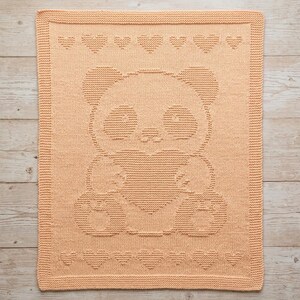 May include: A peach-coloured knitted blanket with a panda bear design. The panda holds a heart, and heart patterns adorn the top and bottom edges. The rectangular blanket has a textured appearance, suitable for a nursery or child's room.