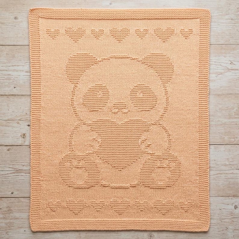 May include: A peach-coloured knitted blanket with a panda bear design. The panda holds a heart, and heart patterns adorn the top and bottom edges. The rectangular blanket has a textured appearance, suitable for a nursery or child's room.