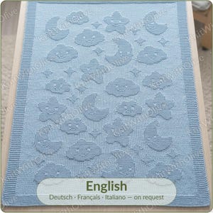 May include: A light blue knitted blanket featuring a repeating pattern of textured, raised motifs including smiling crescent moons, smiling clouds, and five-pointed stars. The blanket has a wide, ribbed border along all four edges.