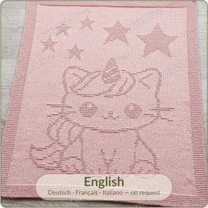 May include: A rectangular light pink knitted blanket featuring a textured pattern of a cat with a unicorn horn on its head. Above the cat, there are five stars of varying sizes. The blanket has a wide, ribbed border along all four edges.