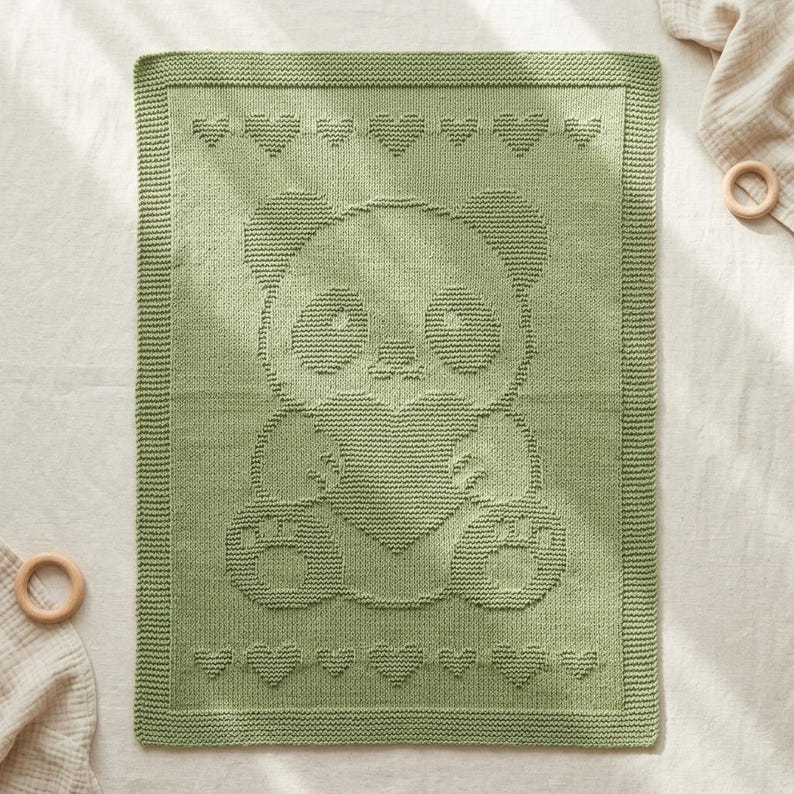 May include: A sage green knitted baby blanket featuring a panda bear holding a heart. The blanket has a border of hearts along the top and bottom edges. The soft blanket is made of a textured knit.
