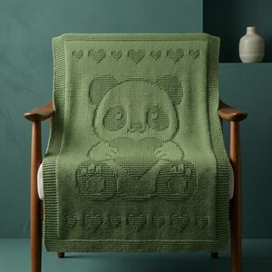 May include: A green knitted blanket with a panda bear design, draped over a wooden chair. The panda is the central focus, surrounded by a border of hearts. The blanket is made of a soft, textured material, perfect for a cosy setting.