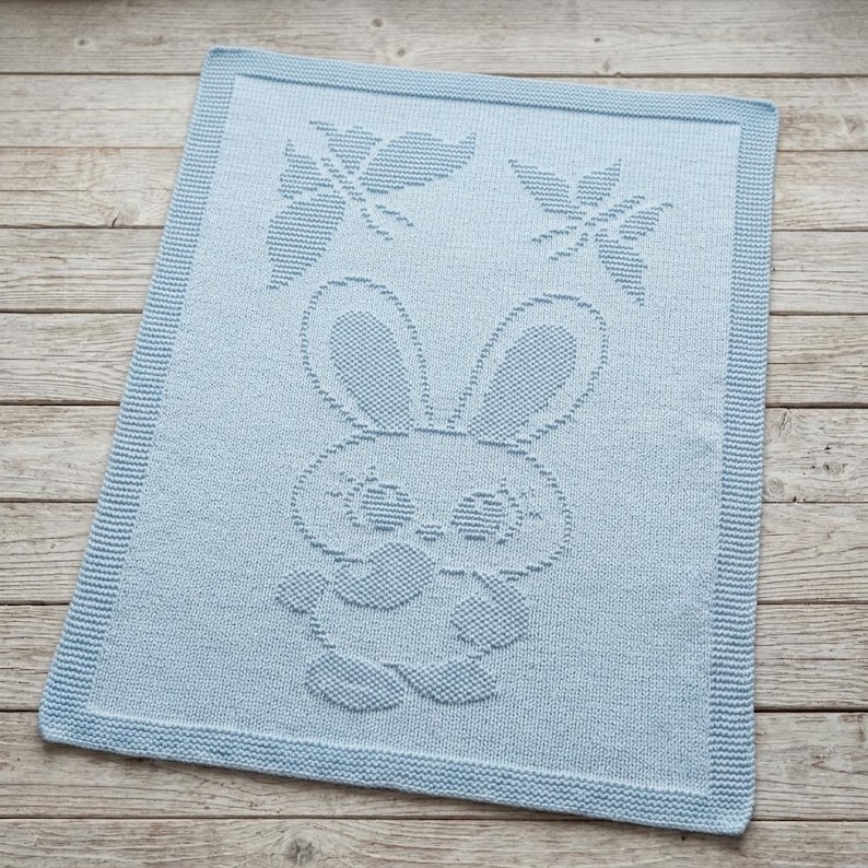 May include: A light blue rectangular knit blanket featuring a textured pattern of a central rabbit character with long ears and two butterflies positioned above the rabbit's head. The blanket has a wide, flat-knit border around the entire edge.