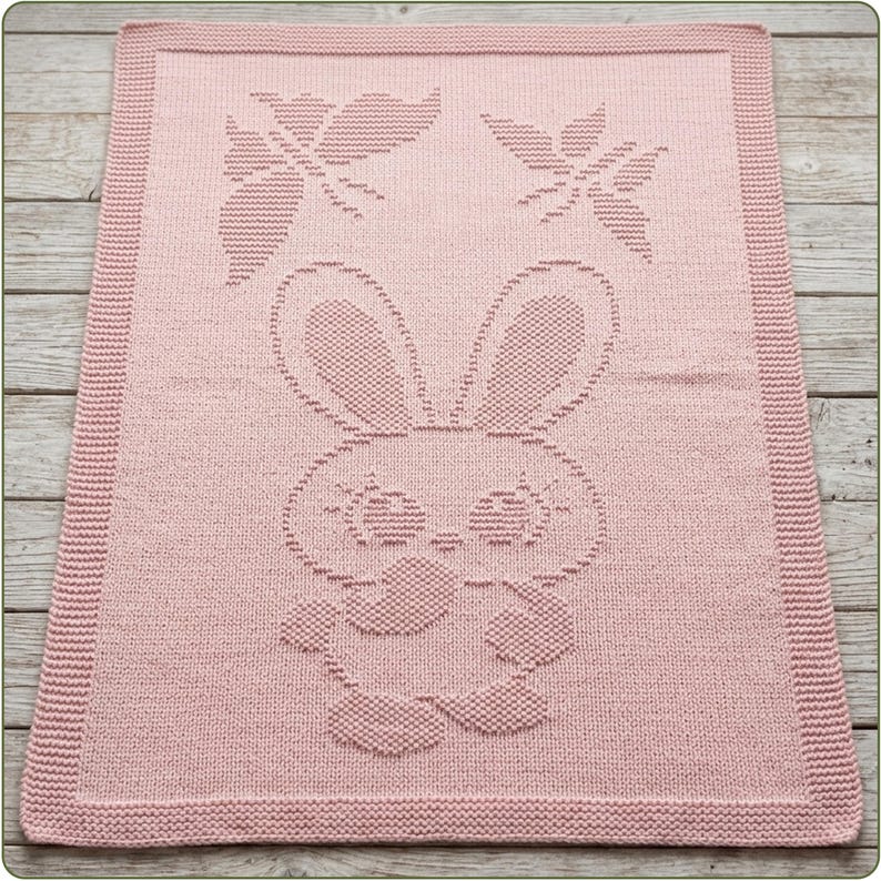 May include: A rectangular light pink knitted blanket featuring a textured pattern of a central rabbit character with large ears and two butterflies positioned above the rabbit. The blanket has a wide, uniform garter stitch border around the edges.