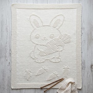 K&ouml;nnte beinhalten: Eine rechteckige, cremewei&szlig;e Strickdecke mit einem strukturierten Muster eines Hasen, der eine Karotte in der Mitte h&auml;lt. Das Design umfasst vier kleinere Karottenmotive am unteren Rand und einen breiten Rand im kraus rechts Muster.