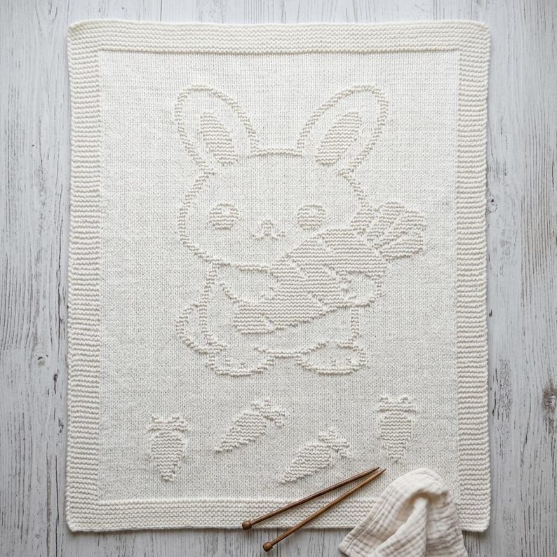 K&ouml;nnte beinhalten: Eine rechteckige, cremewei&szlig;e Strickdecke mit einem strukturierten Muster eines Hasen, der eine Karotte in der Mitte h&auml;lt. Das Design umfasst vier kleinere Karottenmotive am unteren Rand und einen breiten Rand im kraus rechts Muster.