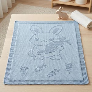 K&ouml;nnte beinhalten: Eine hellblaue Strickdecke mit einem strukturierten Muster aus einem Hasen, der eine Karotte h&auml;lt, umgeben von vier kleineren Karottenmotiven. Die Decke hat einen markanten Rand und ein weiches, gewebtes Erscheinungsbild mit gleichm&auml;&szlig;igem Strick.