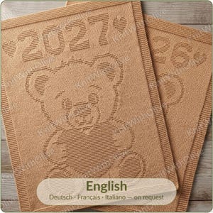 Peut inclure: Deux couvertures tricotées de couleur beige avec un motif d'ours en peluche texturé. La couverture principale affiche l'année 2027 entourée de deux petits cœurs. La seconde couverture partiellement visible montre le nombre 26 et un cœur.