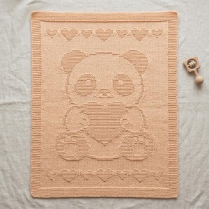 May include: A peach-coloured knitted baby blanket featuring a panda bear holding a heart. The blanket has a border of hearts along the top and bottom edges. A wooden rattle is in the upper right corner.