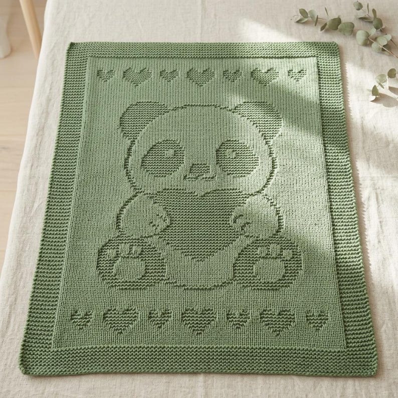 May include: A sage green knitted baby blanket with a panda design holding a heart. The blanket has a border of hearts and a ribbed edge. The panda's face, paws, and heart are detailed with textured stitching.