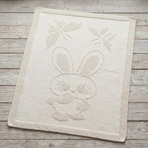May include: A rectangular off-white knit blanket featuring a textured pattern of a central rabbit character with large ears and two butterflies positioned above the rabbit. The blanket has a wide, ribbed border framing the entire design.