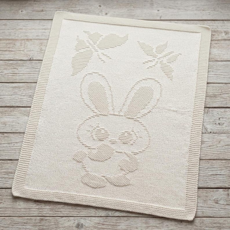 May include: A rectangular off-white knit blanket featuring a textured pattern of a central rabbit character with large ears and two butterflies positioned above the rabbit. The blanket has a wide, ribbed border framing the entire design.