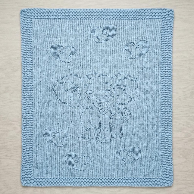May include: A light blue knitted blanket featuring a central elephant motif and five surrounding heart shapes. The blanket has a textured garter stitch border framing the smooth stockinette stitch center where the elephant and hearts are depicted.