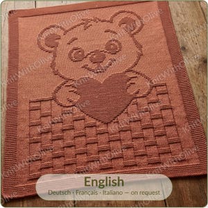 May include: A rectangular terracotta-coloured knitted blanket featuring a textured design of a teddy bear holding a heart while sitting inside a basket. The blanket has a wide garter stitch border and a basket-weave pattern at the bottom section.
