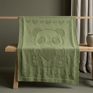 May include: A sage green knitted blanket with a panda bear design. The blanket has a border and a pattern of hearts along the top and bottom. The blanket is draped over a wooden bench.