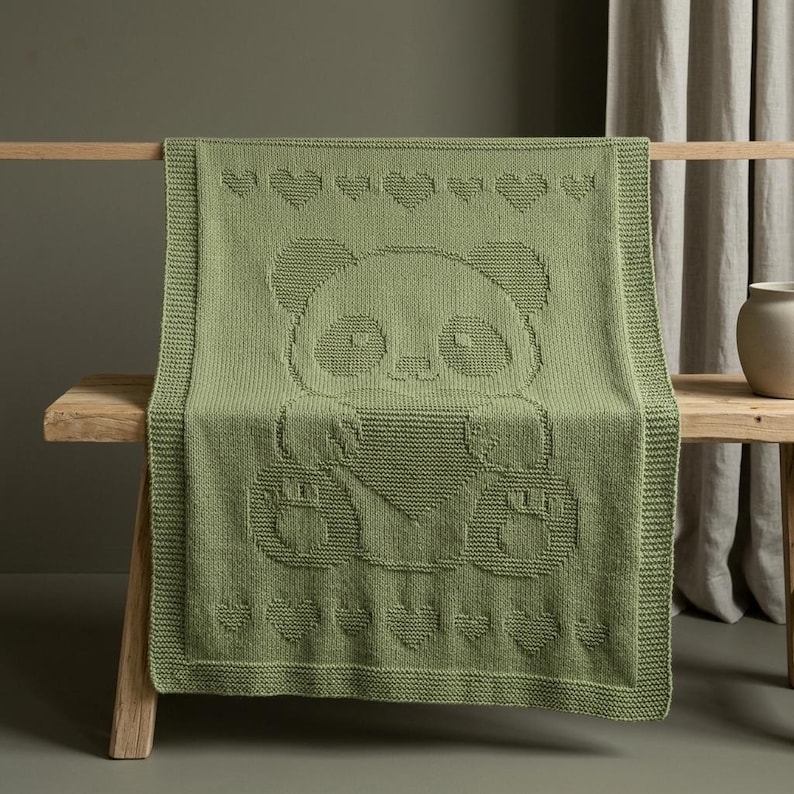 May include: A sage green knitted blanket with a panda bear design. The blanket has a border and a pattern of hearts along the top and bottom. The blanket is draped over a wooden bench.