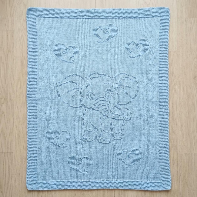 May include: A light blue knitted blanket featuring a textured elephant design in the center, surrounded by five heart motifs. The blanket has a wide, ribbed border that frames the central pattern, creating a consistent look across the entire piece.