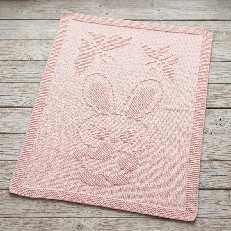 May include: A rectangular light pink knitted blanket featuring a textured pattern of a central rabbit character and two butterflies above it. The blanket has a wide, uniform knitted border framing the central design on all four sides.