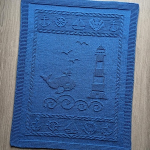Könnte beinhalten: Eine gestrickte blaue Decke mit maritimem Thema. Die Decke zeigt einen Leuchtturm, einen Wal, Möwen und Wellen. Dekorative Bordüren umfassen Anker, Muscheln und Segelboote. Die Decke ist rechteckig.