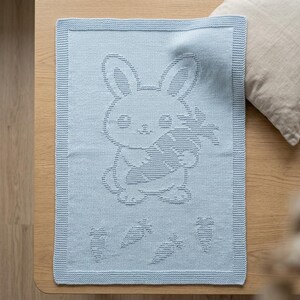 K&ouml;nnte beinhalten: Eine hellblaue, rechteckige Strickdecke mit einem strukturierten, erhabenen Muster eines Hasen, der eine gro&szlig;e Karotte h&auml;lt. Das Design umfasst vier kleinere Karottenmotive unter dem Hasen sowie einen markanten Rand im Kraus-Rechts-Muster.