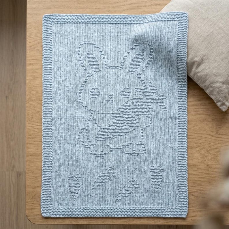 K&ouml;nnte beinhalten: Eine hellblaue, rechteckige Strickdecke mit einem strukturierten, erhabenen Muster eines Hasen, der eine gro&szlig;e Karotte h&auml;lt. Das Design umfasst vier kleinere Karottenmotive unter dem Hasen sowie einen markanten Rand im Kraus-Rechts-Muster.