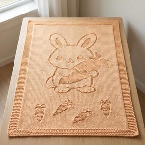 K&ouml;nnte beinhalten: Eine rechteckige, pfirsichfarbene Strickdecke mit einem strukturierten Design eines Hasen, der eine Karotte h&auml;lt. Unter dem Hasen befinden sich vier kleinere Karottenmotive. Die Decke hat einen breiten Rand im Kraus-Rechts-Muster.