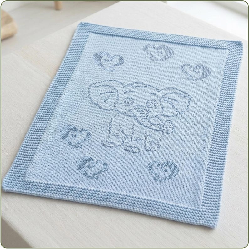 May include: A light blue knitted blanket featuring a central elephant motif surrounded by five stylized heart shapes. The rectangular blanket has a textured garter stitch border framing the smooth stockinette stitch center panel with the design.
